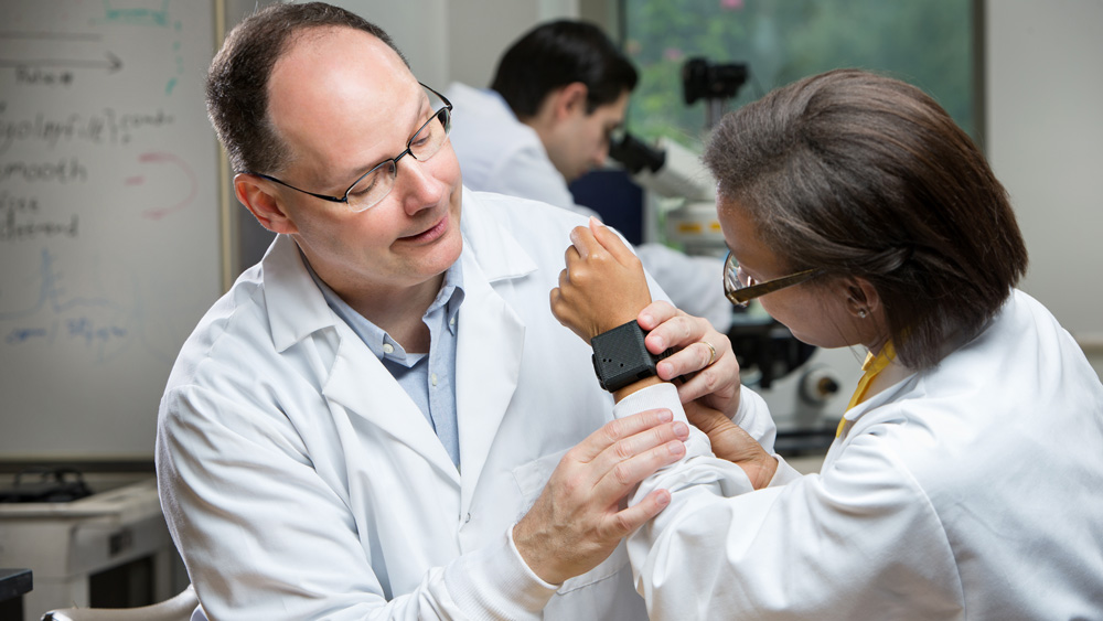 Male professor inspects a black square-shaped medical device on a female student researcher’s wrist