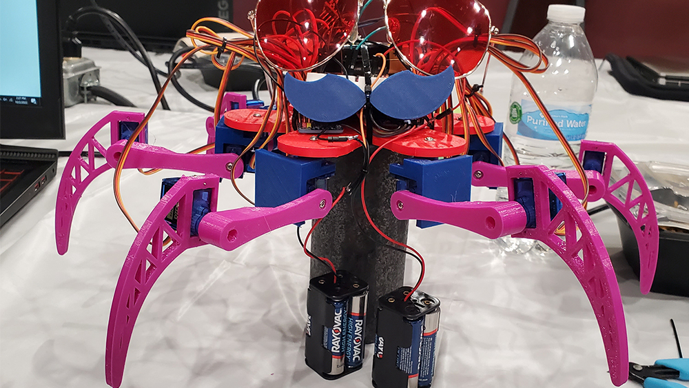 On a table, the six-legged robot Bitwise Boogie wears a mustache and sunglasses with two battery packs attached by wires beneath it.