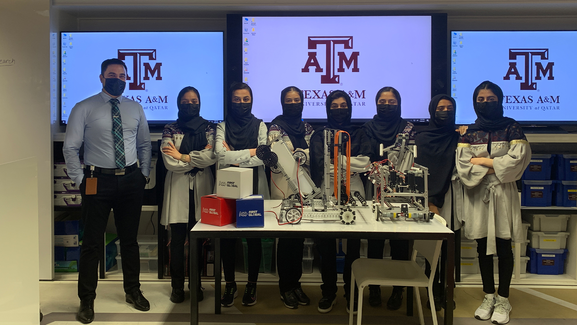 In the foreground, robots sit atop a table. Behind the table, Dr. Mohamed Gharib stands with the Afghan Dreamer team. In the background the Texas A&amp;M University at Qatar logo is displayed on three television monitors.