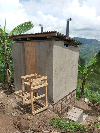 Photo of the latrine that is made with construction materials including wood for the door, medal for the roof, and concrete-like material for walls that sits on rocks on a hill. 