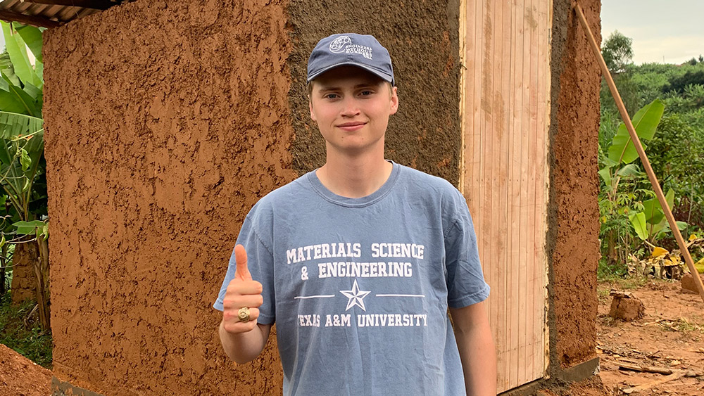 Eli Norris standing in front of the latrine after the build giving a thumbs up, wearing a blue shirt that says “Material Science and Engineering, Texas A&amp;M University