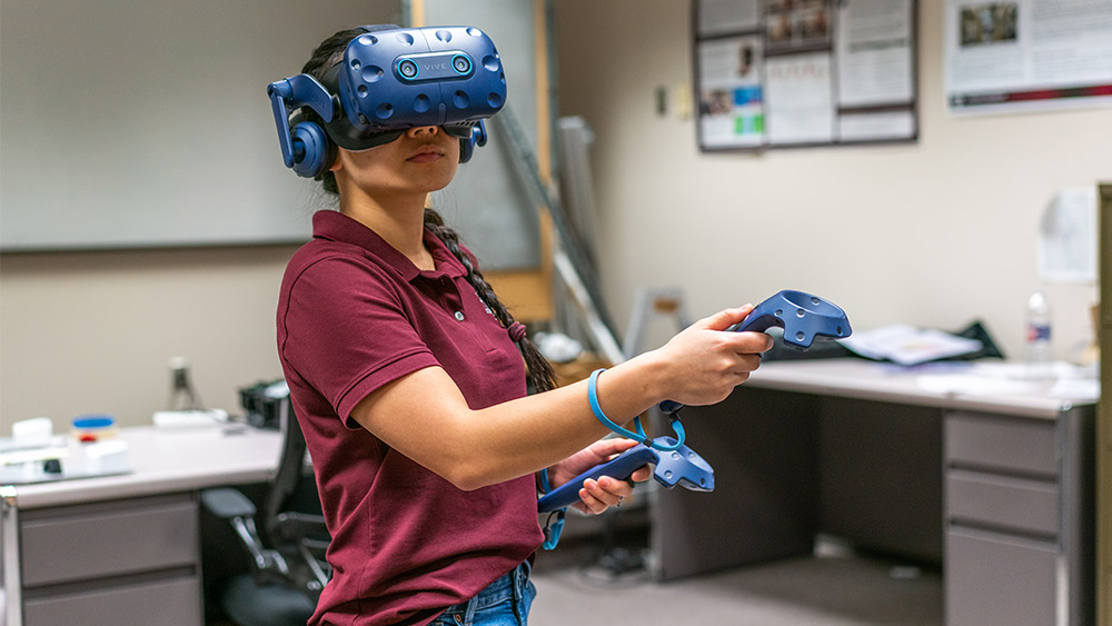 A female student wearing a VR headset and holding controls in a research laboratory.