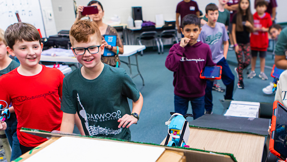 Students stand around a cardboard ramp as they observe a wheeled robot climb the ramp.