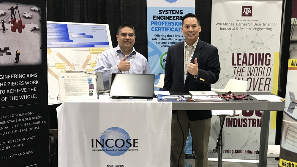 Two men stand behind a table at an INCOSE conference. Around them are posters with the industrial and systems engineering logo and the INCOSE logo.