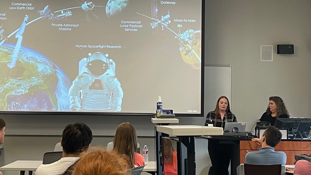 A female researcher stands at the front of a classroom giving a presentation. On the projector screen is an image of an astronaut in space with words “human space flight research.”