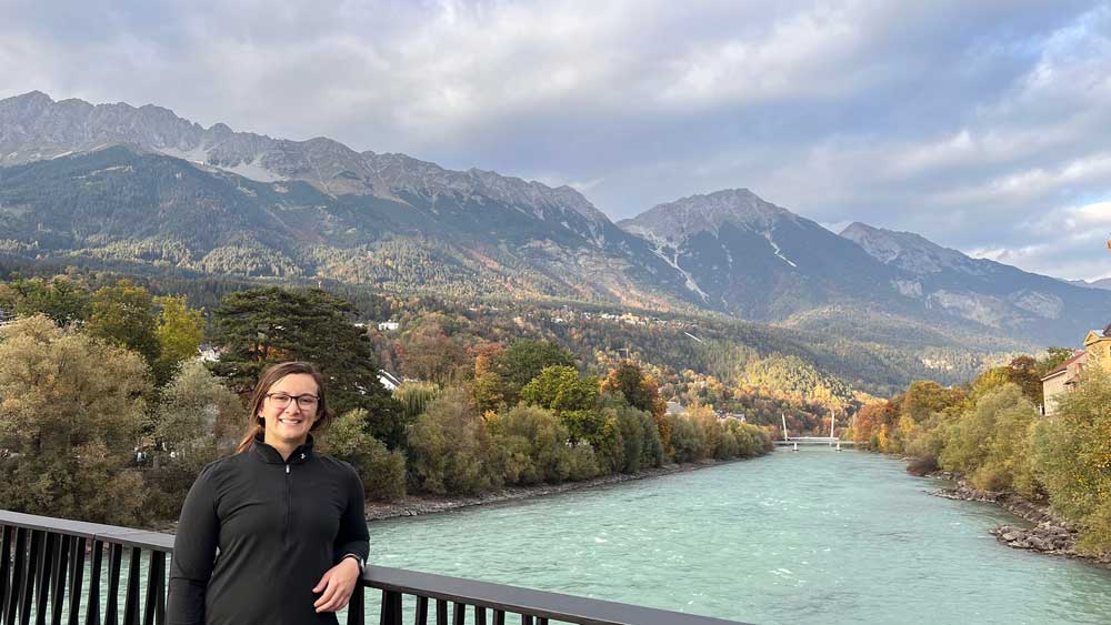 Whitney Mantooth stands on a bridge above a river in Tyrol, Austria. Behind her is fall foliage and mountains. 