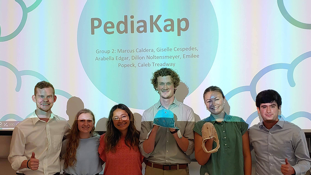 Group of former biomedical students standing in front of a screen that reads, “PediaKap.” Two of the students hold caps.