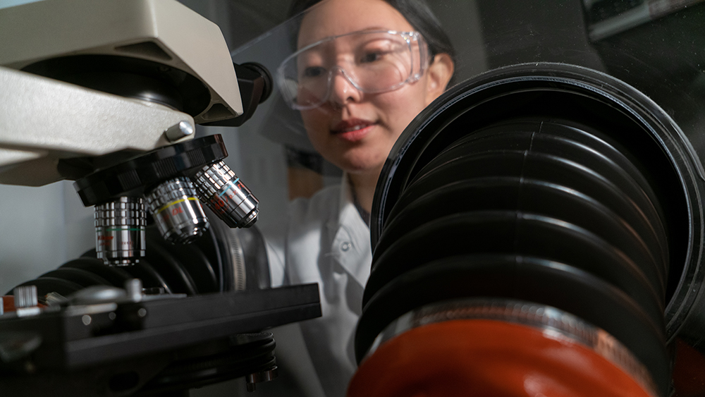 A female researcher wearing safety glasses and a lab coat adjusts the focus on a complex microscope.