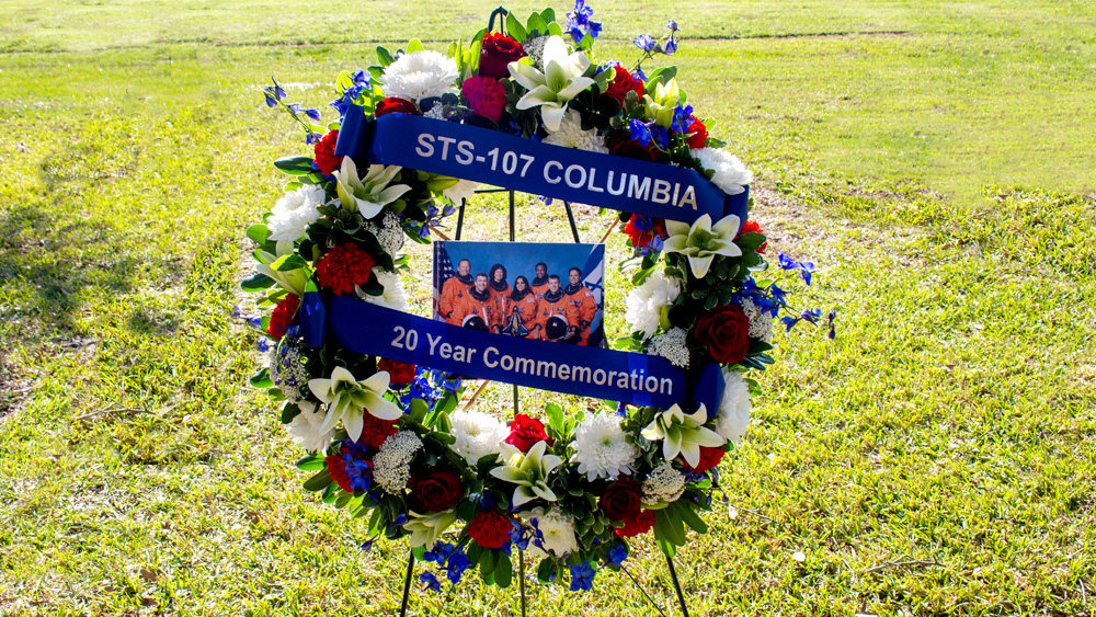 A wreath with flowers stands in a park. In the middle of the wreath is a photo of seven astronauts. A blue ribbon reads “STS-107 Columbia 20 year commemoration.”
