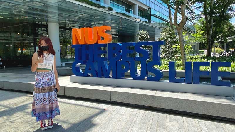 Mia Nguyen stands outside her dorm building on the National University of Singapore campus by a sculpture sign that reads “NUS: The Best Campus Life.”