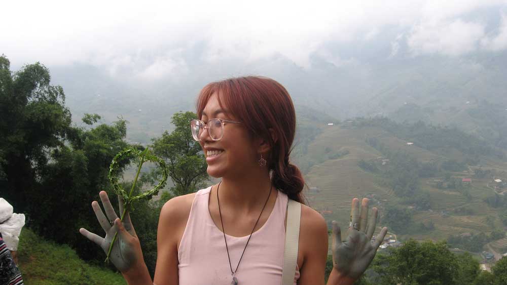 Mia Nguyen stands on a mountain, hands covered in natural indigo dye. Behind her are mist-covered mountains and a green valley dotted with housing structures. 