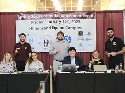 Several smiling college students seated and standing behind check-in tables. Text on sign behind them reads, “Spring 2023 Career Enhancement Event, Friday, February 10th, 2023, Hildebrand Equine Complex.”