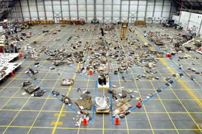 A large floor is covered in pieces of metal, plastic and more materials. Items are placed in a shuttle shape following tape markers on the ground.