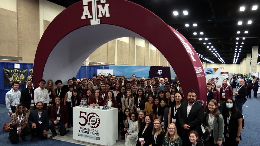 Faculty, staff and students from the Department of Biomedical Engineering at Texas A&M University crowd around the department’s space in the Biomedical Engineering Society exhibit hall in front of the Texas A&M branded maroon arch.