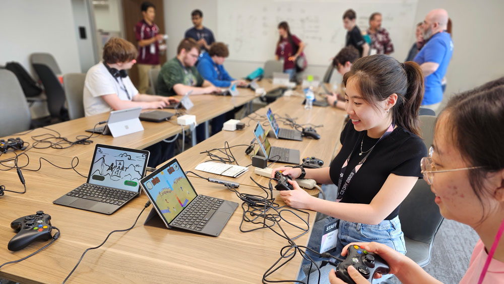 Students sitting at a grouping of tables, using controllers to play video games on a Microsoft Surface tablet.