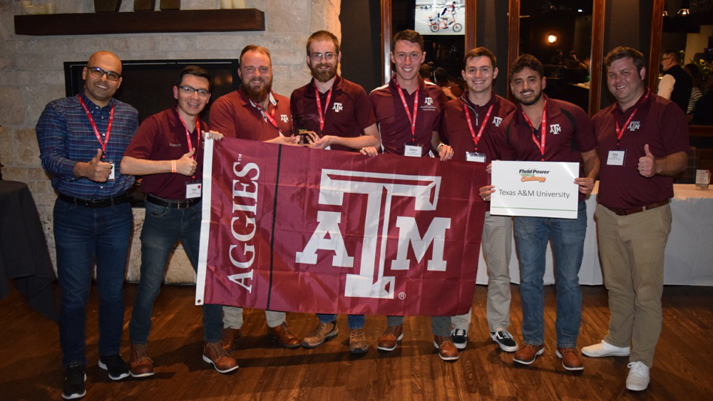 The team — including, from left: Dr. Ahmed Abdelaal, Ivan Franco, Aquiles Alonso, Mark Finley, Dalton Yingst, Preston Baumgartner, David Johnson, Gary Bradley — with flag featuring Aggies and the Texas A&amp;M University logo. Sign reads: Fluid Power Vehicle Challenge, Texas A&amp;M University.
