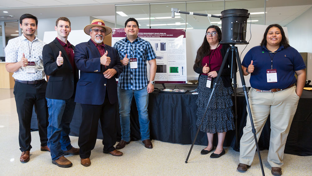 Left to right: William Astorga, Nathaniel Bauer, Professor Oscar Lopez, Omar Tapia, Miriam Alanis, and Katia Melo give thumbs up next to their presentation board and a bucket mounted on a tripod at the Engineering Showcase.