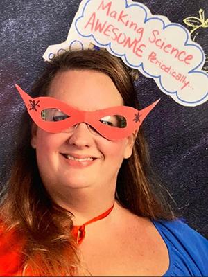 A woman wears a red superhero mask, red cape and blue shirt as she stands against a chalkboard background with a paper thought bubble with the words “Making Science AWSOME periodically.”