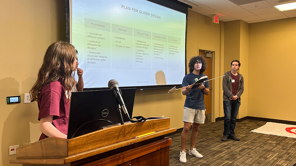 Four students stand in front of a project screen to present and showcase the design of their glider.