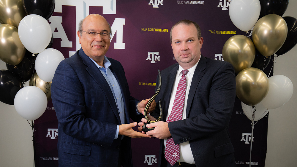 Standing in front of a backdrop with a Texas A&amp;M logo, Dr. Dimitris Lagoudas hands an award to Jason Henderson.