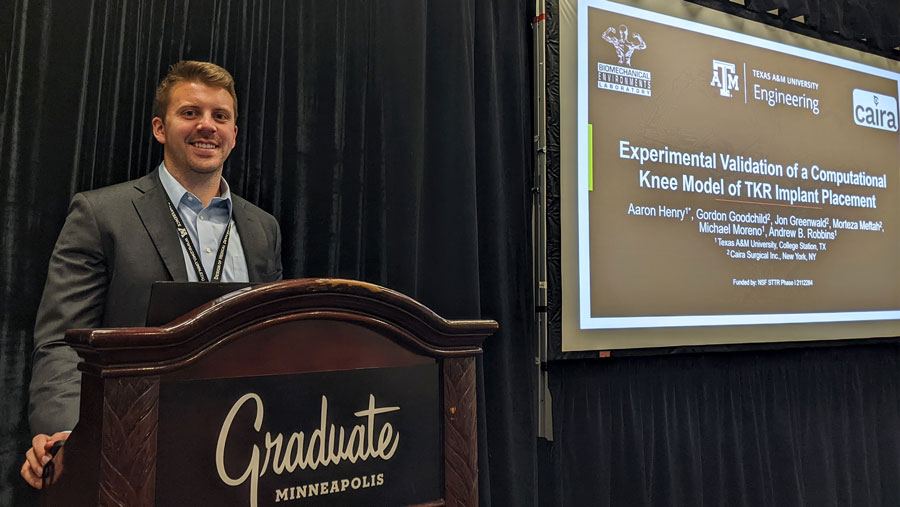 Henry stands at a podium in business attire next to a screen stating the title of his research paper, “Experimental Validation of a Computational Knee Model of TKR Implant Placement.”
