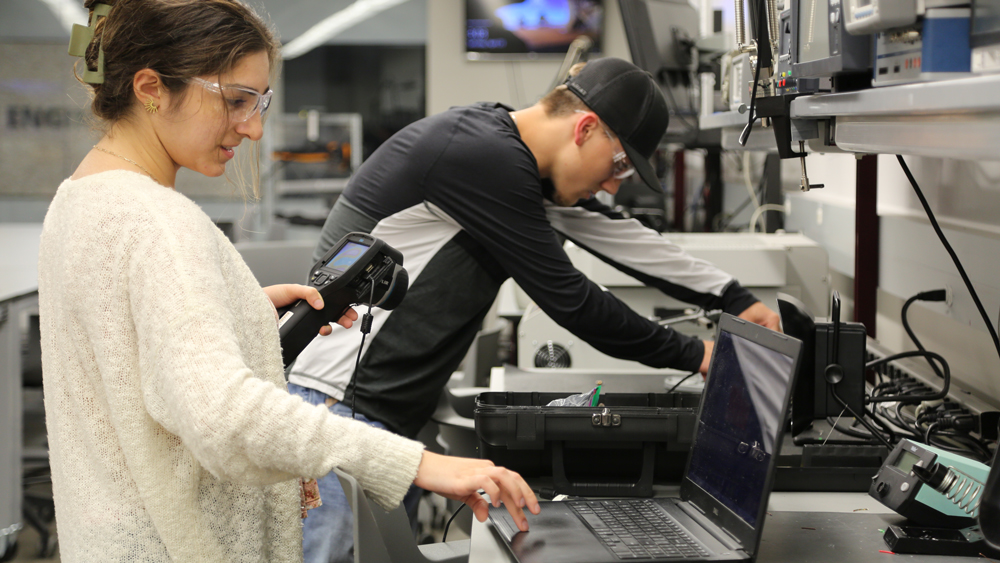 A female student and a male student work on prototypes in the Fischer '72 Engineering Design Center. The female student is holding a thermal device while coding on a laptop.