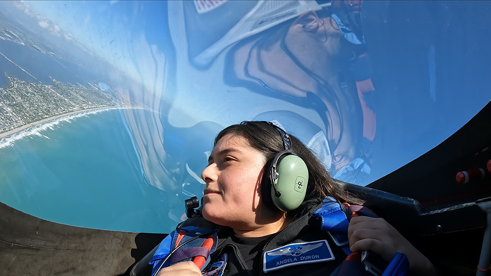 A female seated in the cockpit of a vehicle in flight with water, land and sky visible through the clear cockpit cover behind and above her head.