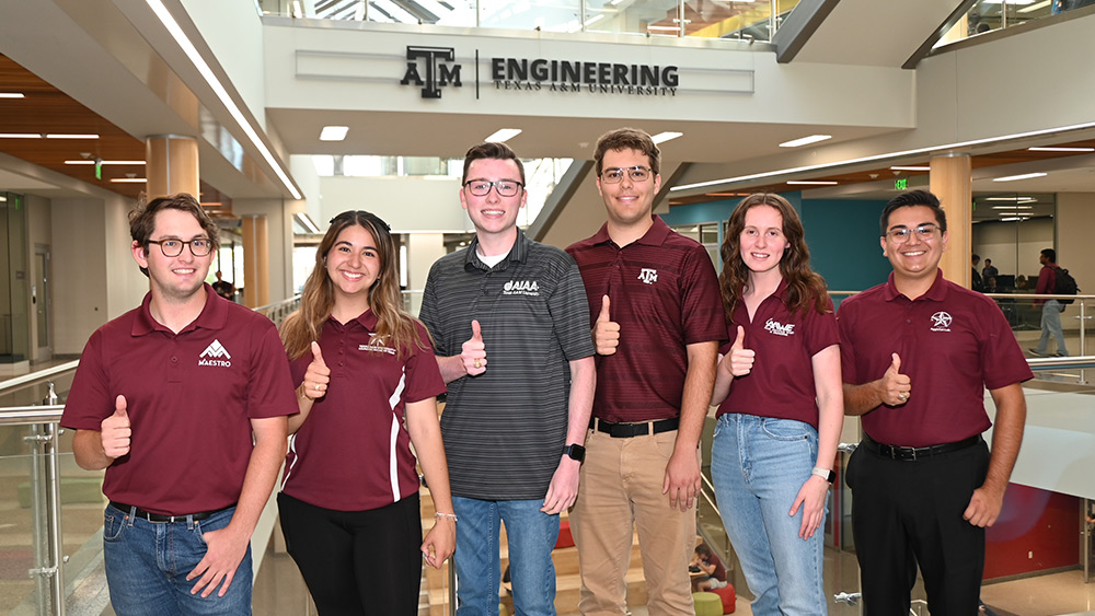 Six students in Texas A&amp;M branded polos smiling and giving a Gig ‘em in the Zachry Engineering Education Complex.