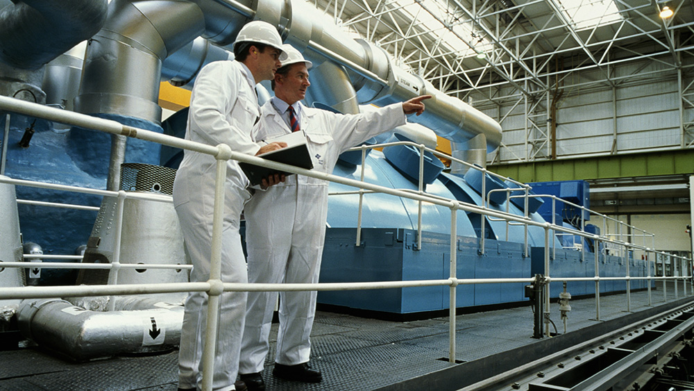 Two men in white safety suits and hard hats perform safety checks at nuclear reactor. 