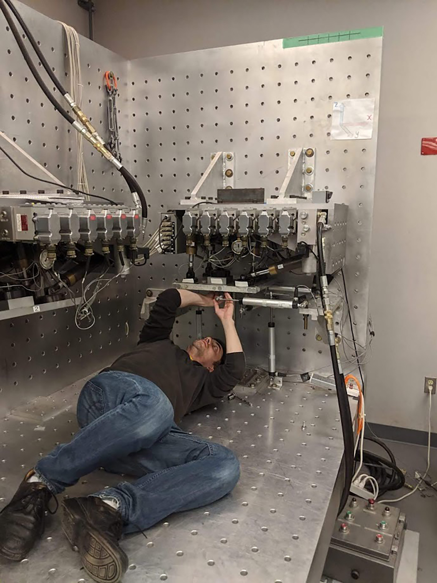 Najafi lays on his back under a testing unit, adjusting an experimental setup with tools lying around him.