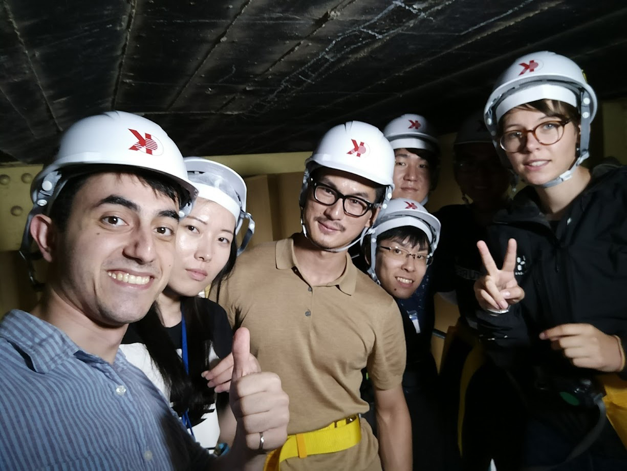 Najafi poses with colleagues in a group photo. Everyone is wearing white hard hats, smiling and standing near a large steel beam in the background.