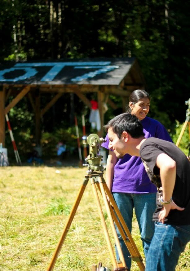 Najafi looks through survey equipment mounted on a tripod in a green grass field around other camp students.
