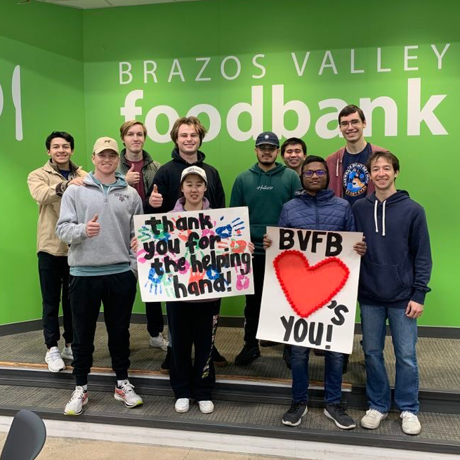 A group photo of the HKN members volunteering at the Brazos Valley Food Bank and holding posters that read "thank you for the helping hand" and "BVFB loves you."