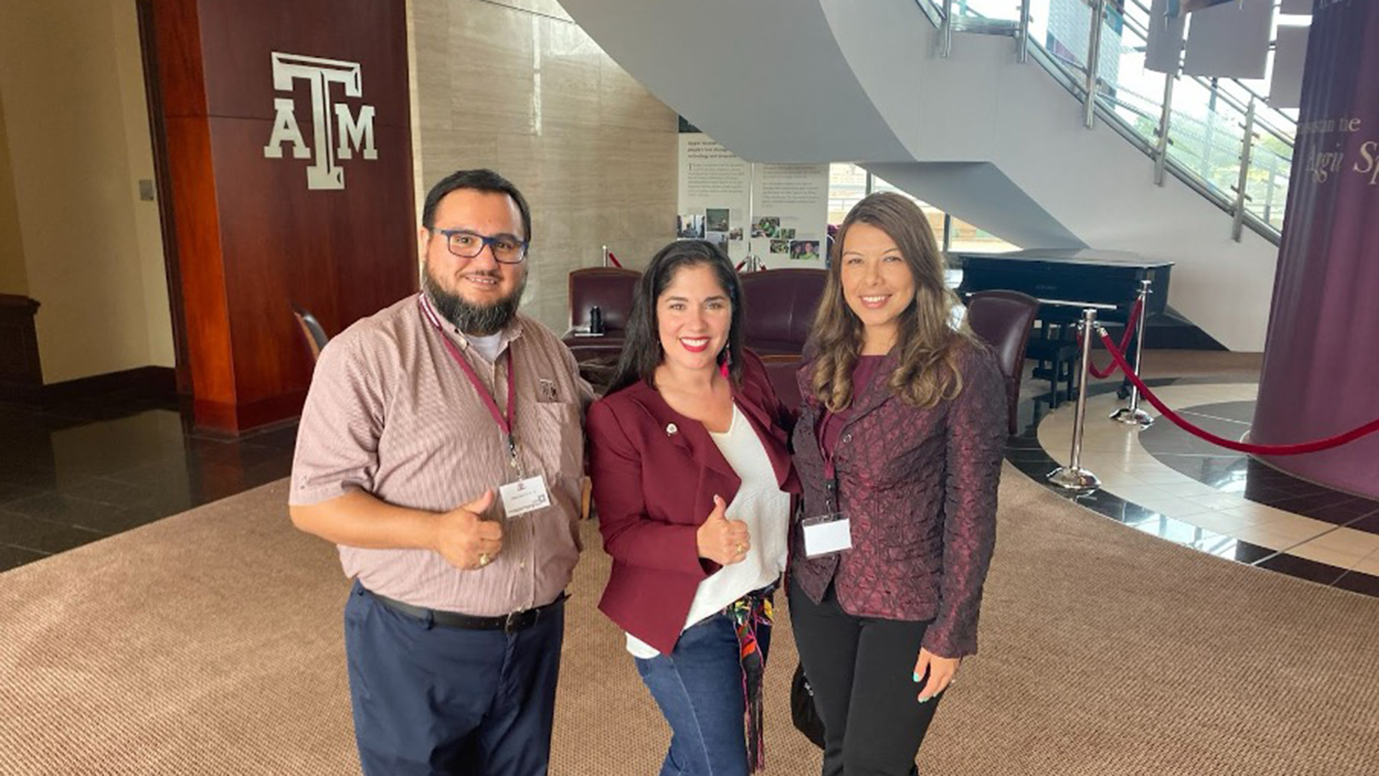 Dr. Jennifer Carvajal in an academic building with Mario Hernandez and Tamara Cuellar, two leaders at a former student association, who are giving a thumbs up with the A&amp;M logo in the back. 