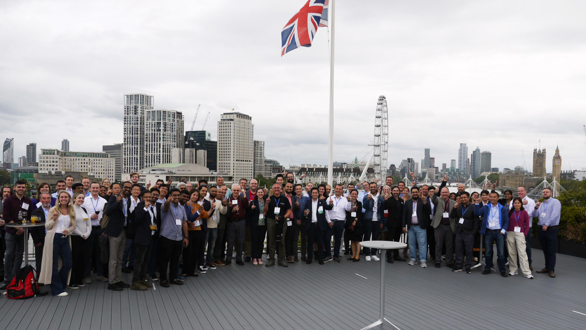 A large group standing on a balcony with the London skyline and London Eye in the background with an English flag waving.