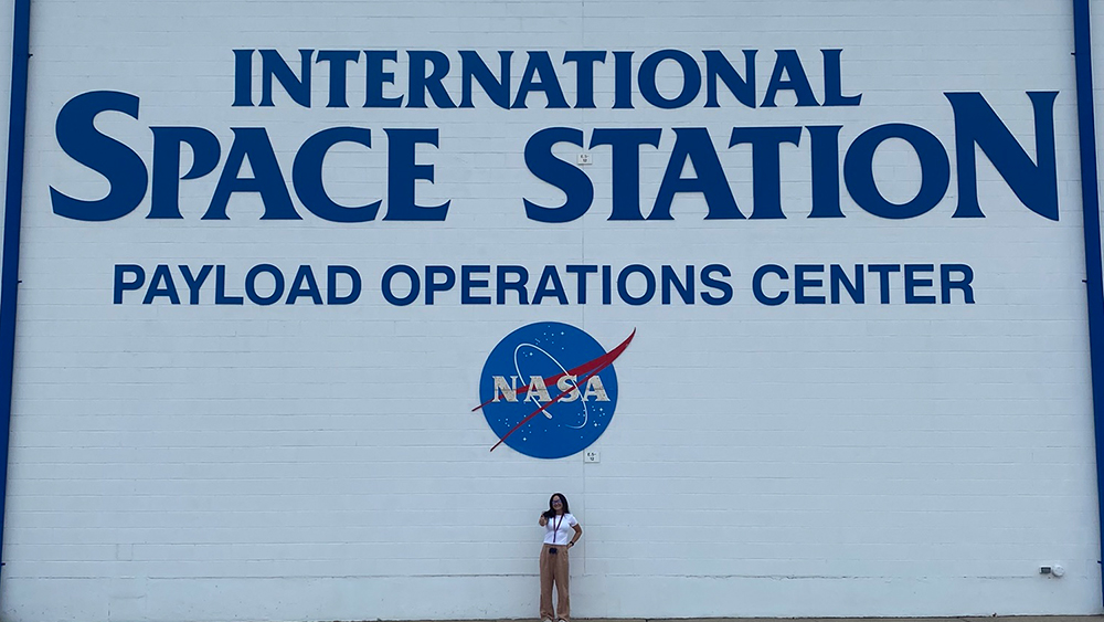 Sofi English wearing white shirt and tan pants stands in front of large white building wall with the words “International Space Station Payload Operations Center” in blue font and the NASA logo on the building. 