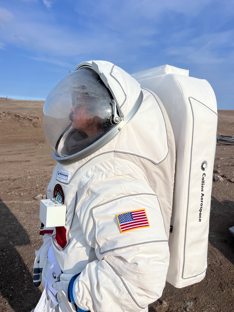Sofi English stands outside on rocky ground wearing white space suit with face shield and backpack like pack. 