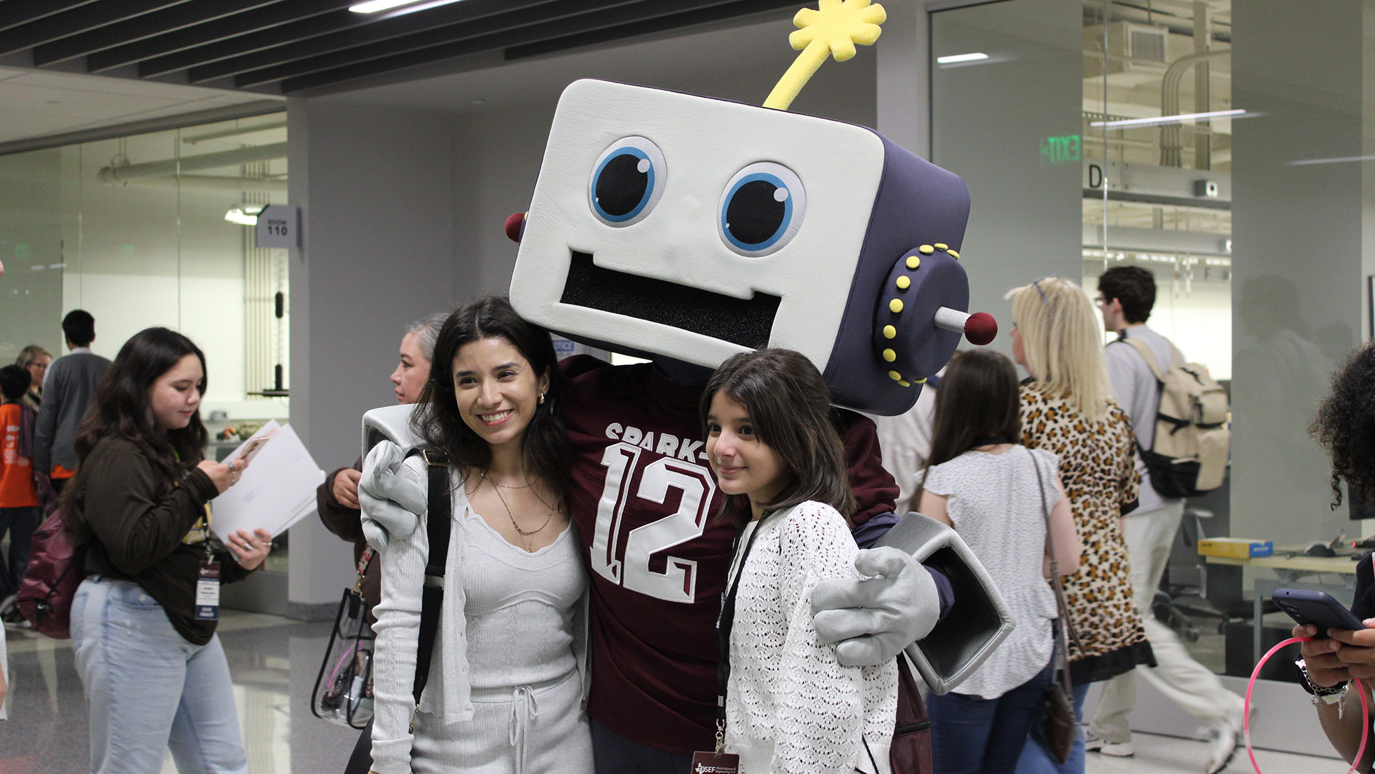 Two people posing for a photo with a person in a large, cartoonish robot costume at an indoor event.