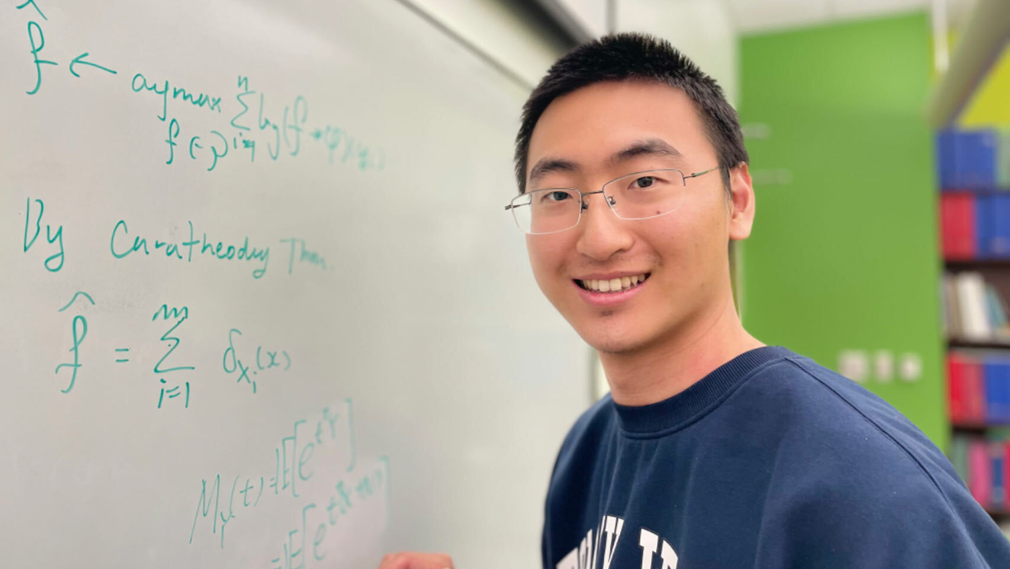 A man in glasses smiling at the camera while writing mathematical equations on a whiteboard in a classroom setting.