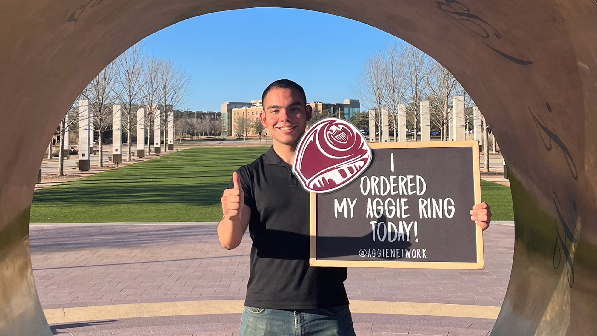 Man poses under Aggie Ring replica with a sign that reads "I ordered my Aggie ring today!"