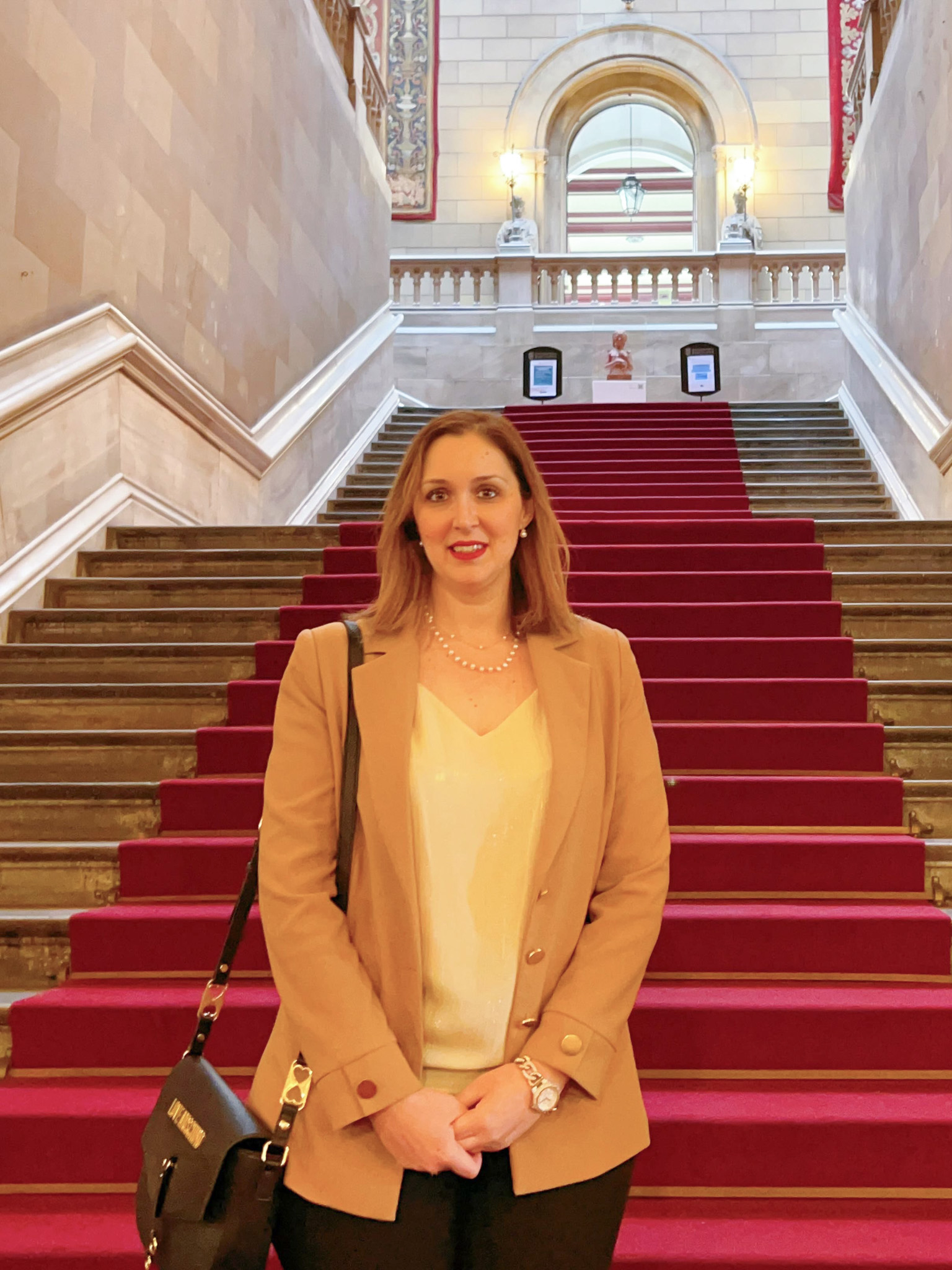 A woman stands on the grand stairway of a historic building.