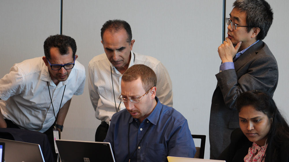 Male and female sit around table working on laptops with three males looking over shoulders.