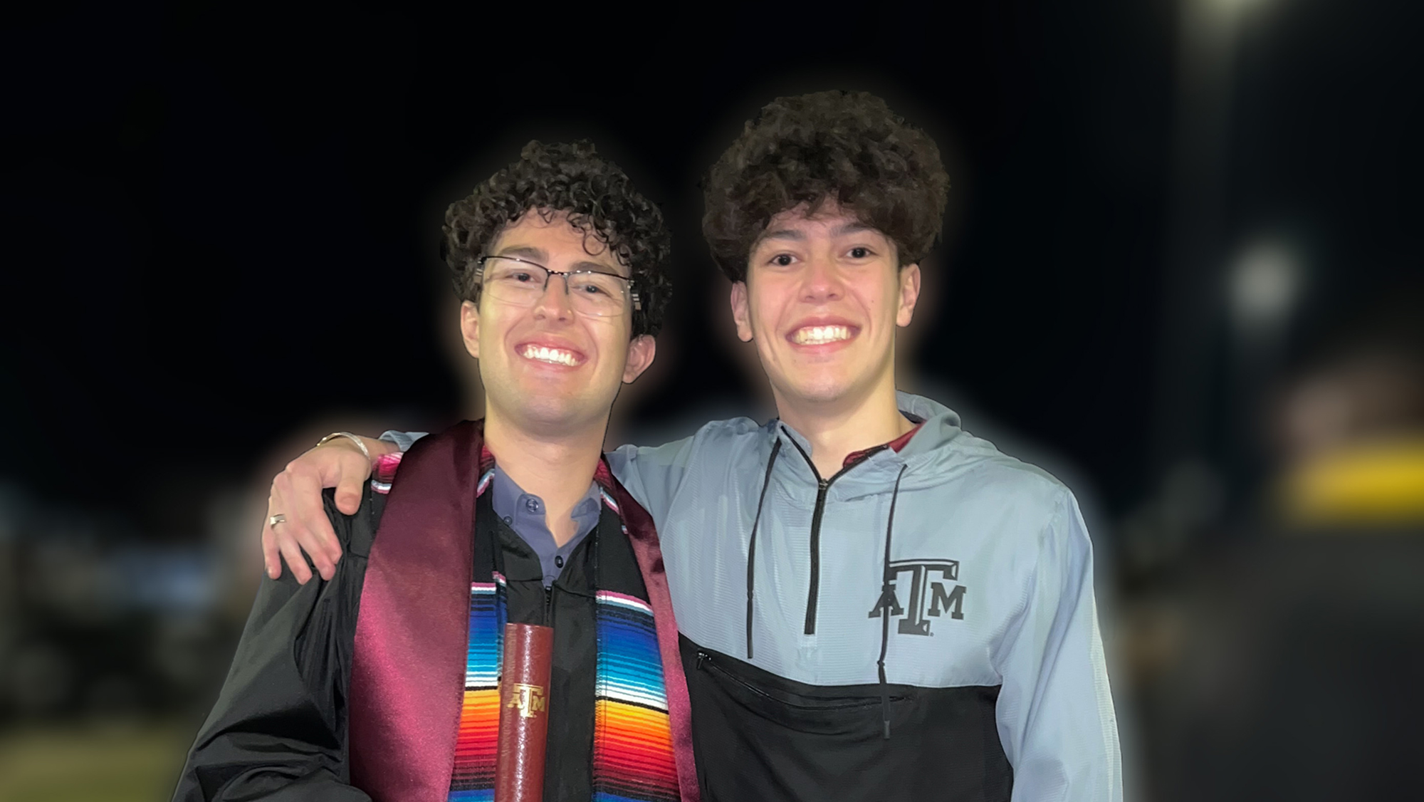Two young men, one wearing graduation regalia, stand with their arms around each other. 