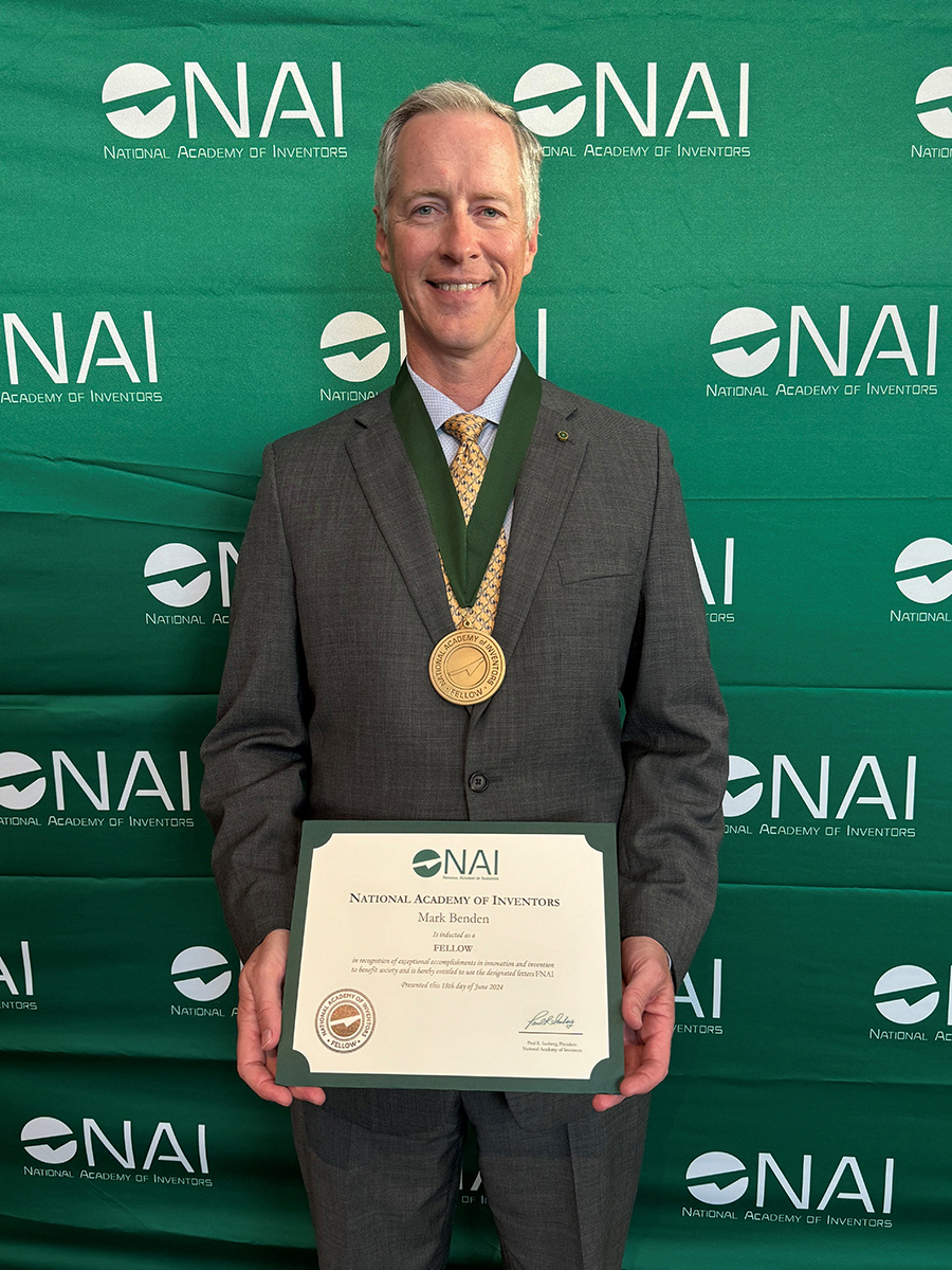 A man is wearing a medal and holding a certificate in front of a banner with the National Academy of Inventors logo.