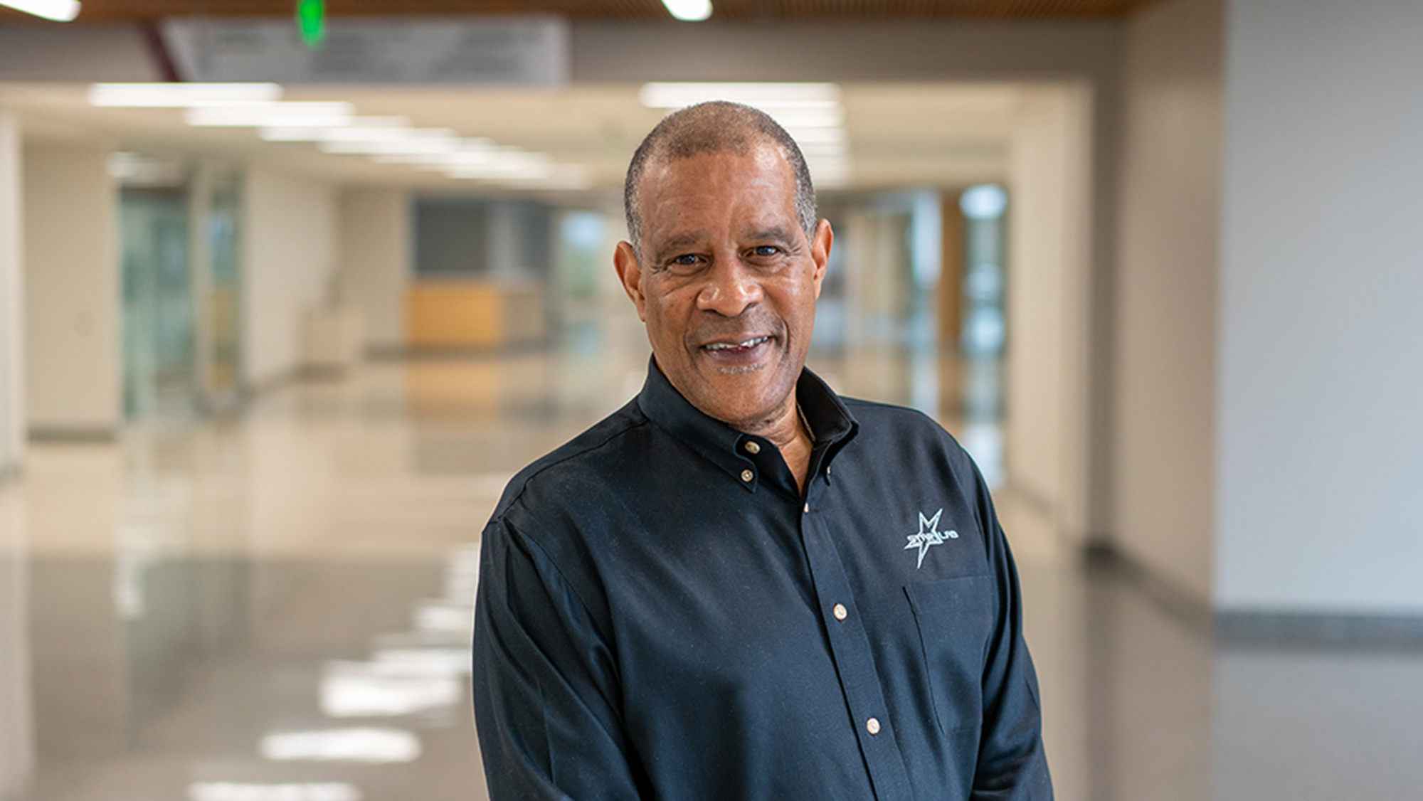 A man with a warm smile standing in the hallway of an office building or institution.