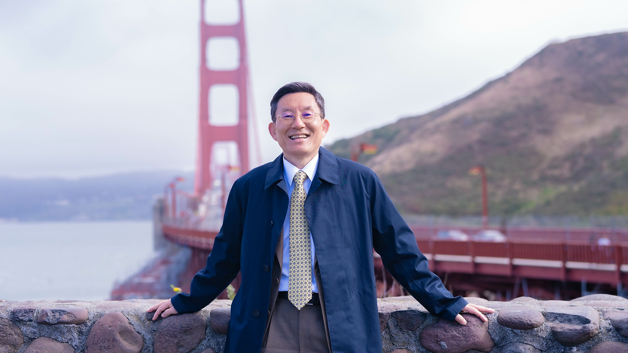 A man in business attire smiling in front of a bridge.