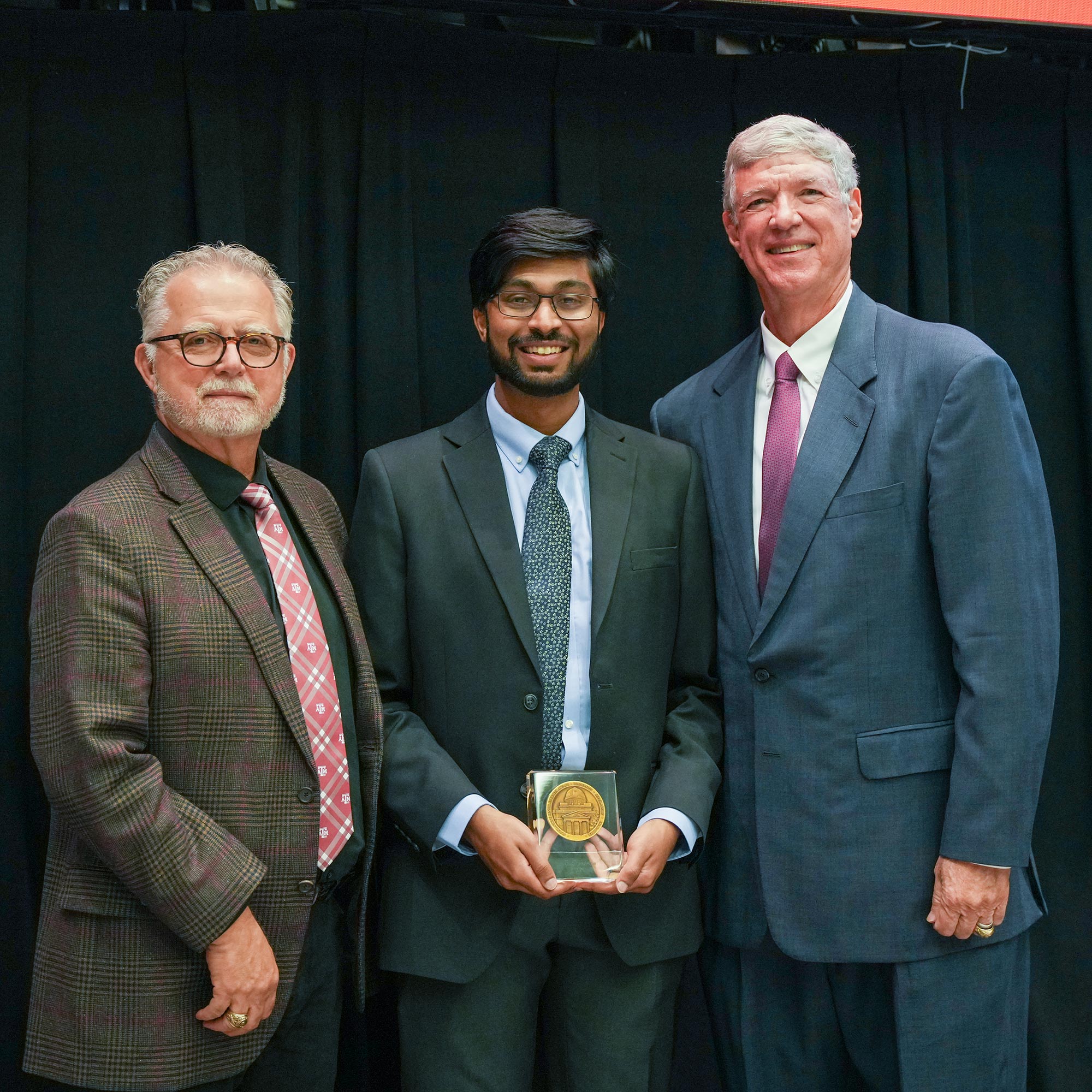 Three men standing, one with an award.