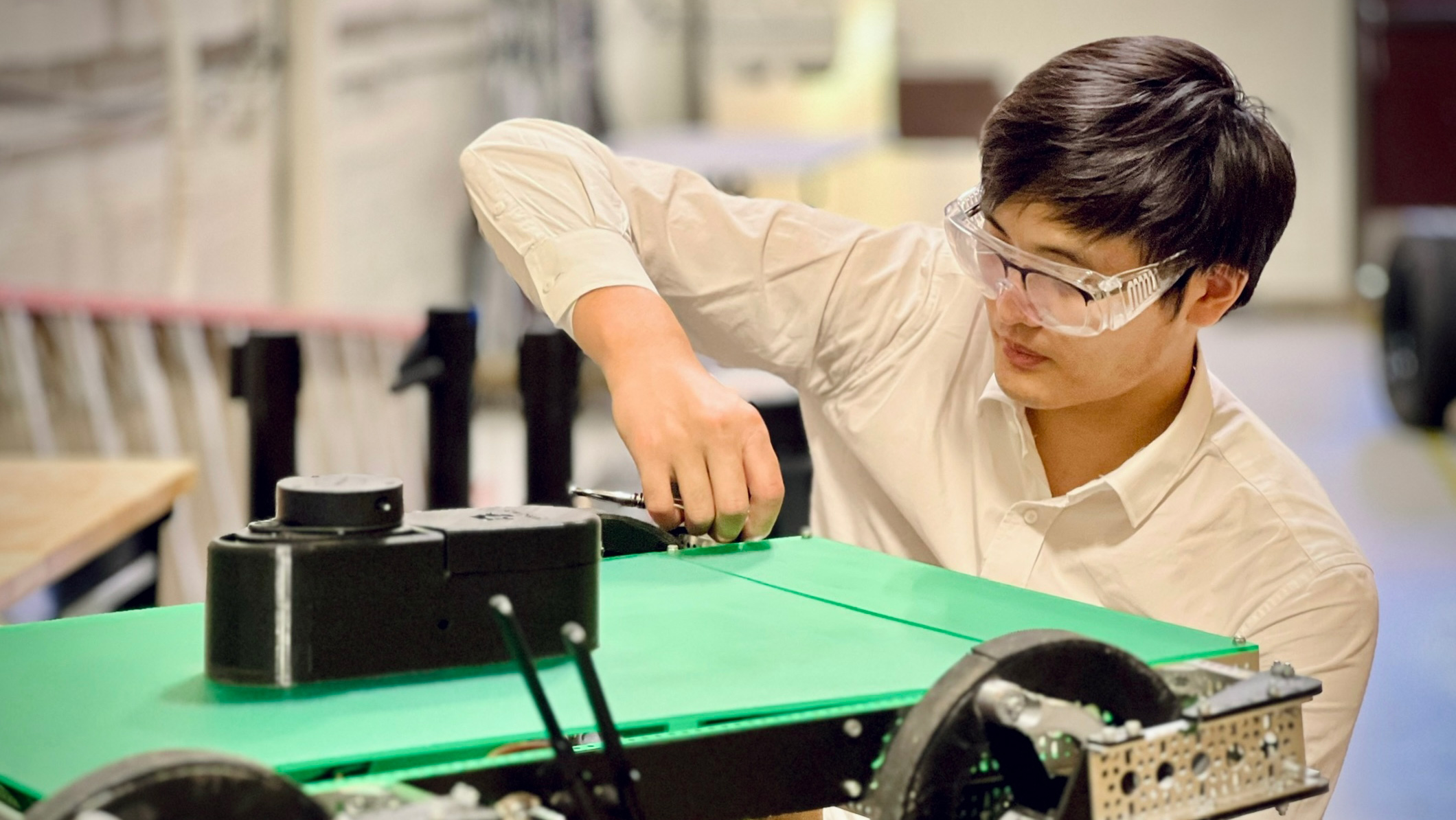 A man with short, dark hair uses a tool to work on the wheel of a green and black robot.