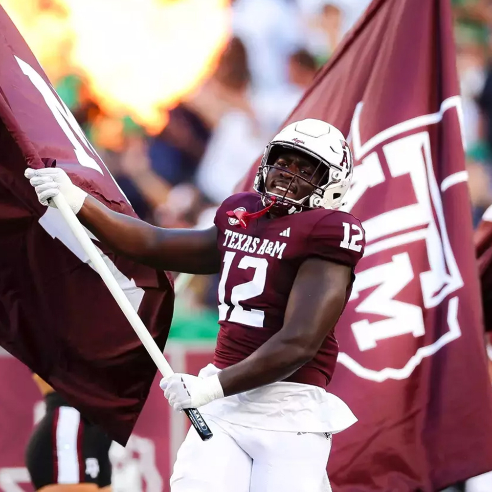 A football player in a maroon jersey waving a flag runs on a football field.