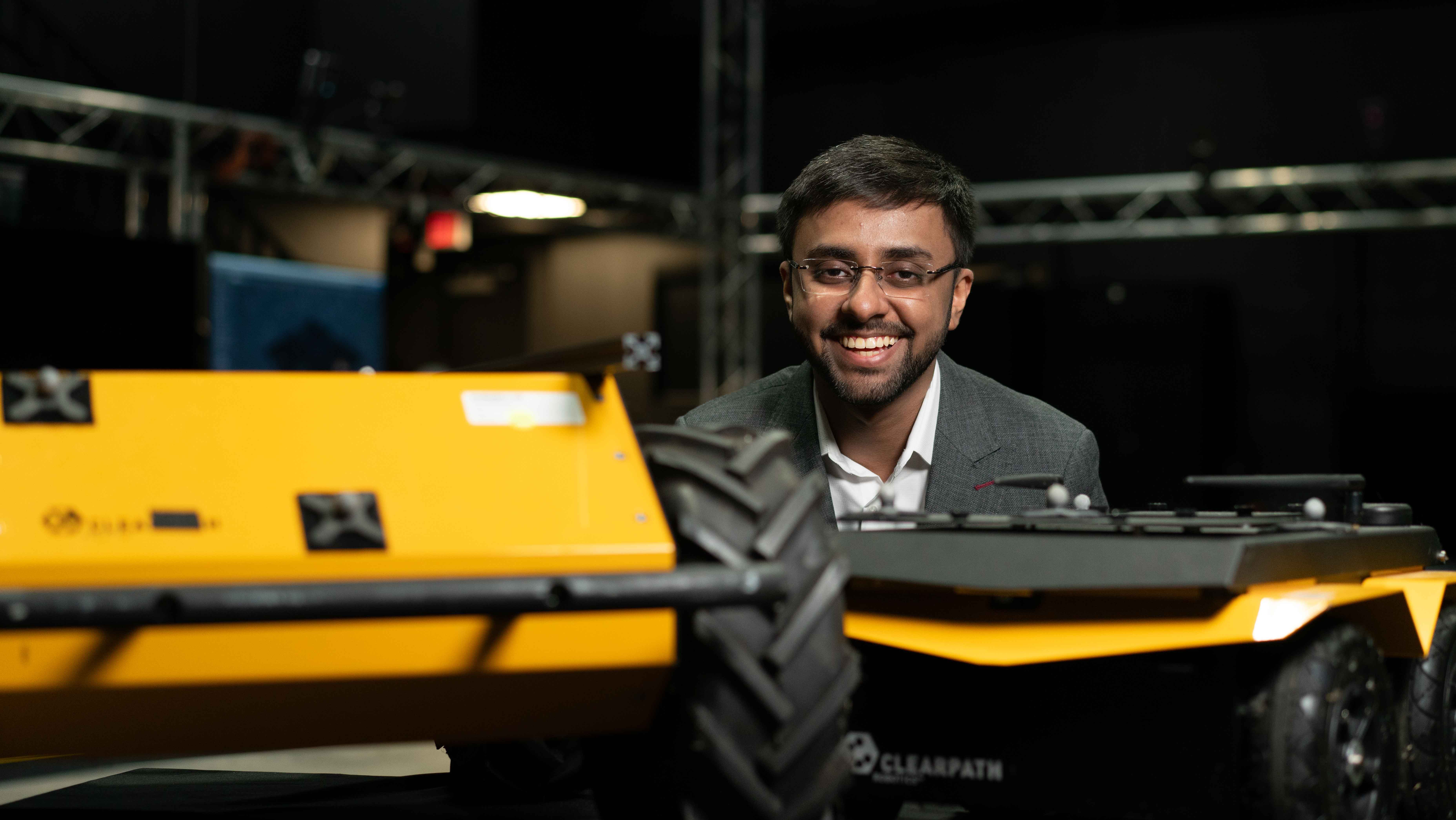 A smiling man in a suit is positioned next to a yellow robotic vehicle.